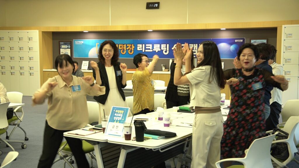 삼성생명 FC 대상 조직 활성화 팀빌딩 행사에서, 수십 명의 여성 영업사원들이 활짝 웃으며 손을 흔들거나 박수를 치고 있습니다. 참가자들의 밝은 표정과 활기찬 모습에서 매우 흥겨운 현장 분위기를 느낄 수 있습니다. 전면의 플래카드와 여러 테이블, 참가자들의 이름표가 보입니다.