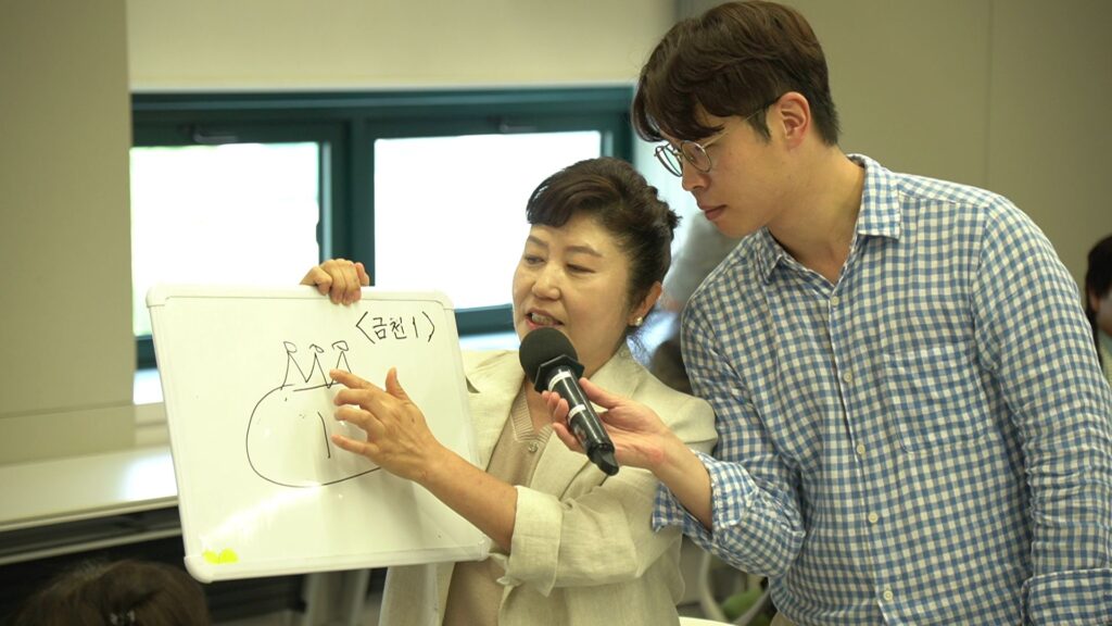 삼성생명 FC 대상 조직 활성화 팀빌딩 행사 중, '금천 1조'의 여성 영업사원이 미니 화이트보드에 직접 그린 산 모양의 그림을 열정적으로 설명하고 있습니다. 남성 전문 MC가 옆에서 마이크를 들고 FC의 발표를 보조하며, FC는 그림의 특정 부분을 손가락으로 가리키고 있습니다. 화이트보드에는 한글로 '<금천 1>'이라고 적혀 있습니다.