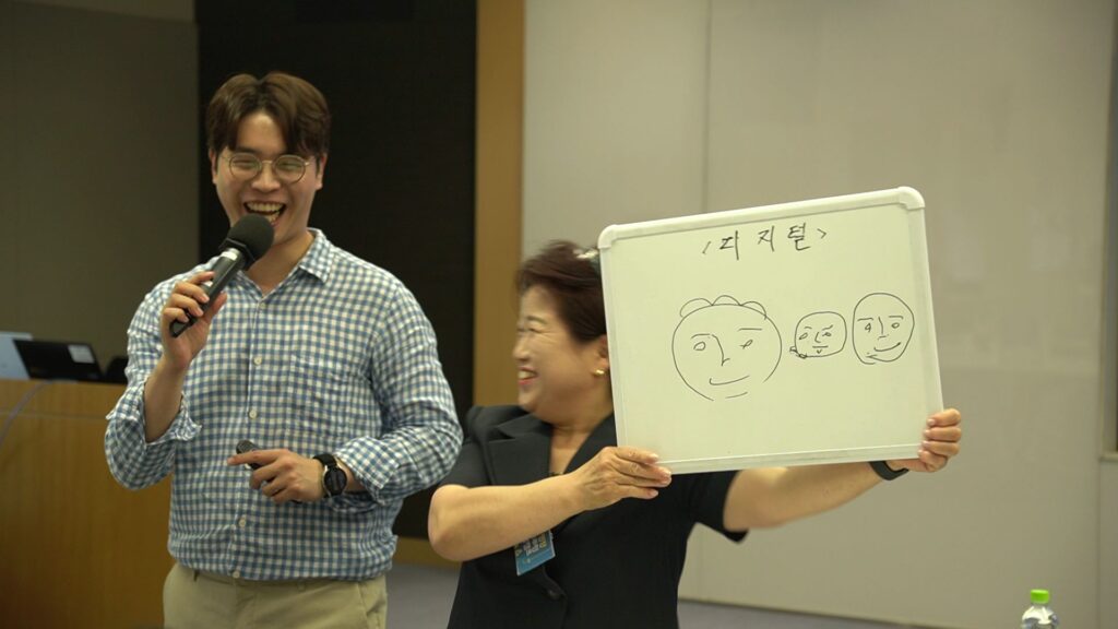 삼성생명 FC 대상 조직 활성화 팀빌딩 행사에서, '디지털'조의 여성 영업사원이 미니 화이트보드에 그린 세 개의 다른 얼굴 표정 그림을 보여주며 활짝 웃고 있습니다. 그 옆에서 남성 전문 MC가 마이크를 들고 함께 즐겁게 웃고 있으며, 두 사람의 밝은 표정에서 활기찬 현장 분위기를 느낄 수 있습니다. 화이트보드에는 한글로 '<디지털>'이라고 적혀 있습니다.