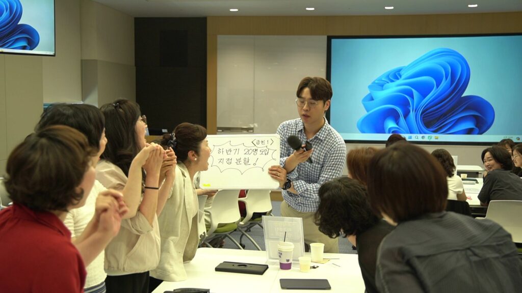 삼성생명 FC 대상 조직 활성화 팀빌딩 행사에서, 남성 전문 MC가 마이크를 들고 미니 화이트보드를 보여주며 설명하고 있습니다. 화이트보드에는 한글로 '<금천 1>', '하반기 20명 도입 지점 분할!'이라고 적혀 있습니다. 여러 명의 여성 영업사원들이 MC의 설명을 경청하며 손을 맞잡고 협동하는 모습을 보여줍니다.