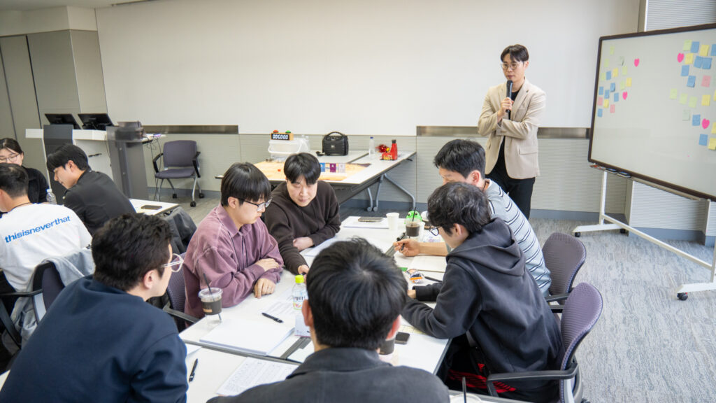 사내 워크샵 실내 프로그램 중 팀원 각자의 강점을 살려 역할 분담을 정하고 보드게임 전략을 치열하게 논의하는 기업 임직원들