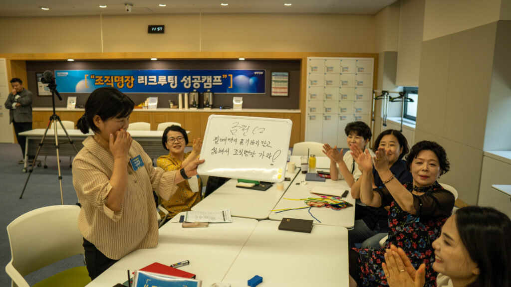 삼성생명 FC 대상 조직 활성화 팀빌딩 행사 중, '금천 C2' 조의 여성 영업사원들이 "될 때까지 포기하지 말고 실행해서 조직명장 가자!"라는 팀의 목표와 다짐이 적힌 미니 화이트보드를 들고 발표하고 있습니다. 같은 조원들이 환하게 웃으며 박수를 치고 응원하는 모습에서 끈끈한 팀워크가 돋보이며, 배경에는 "조직명장 리크루팅 성공캠프" 현수막이 걸려 있습니다.